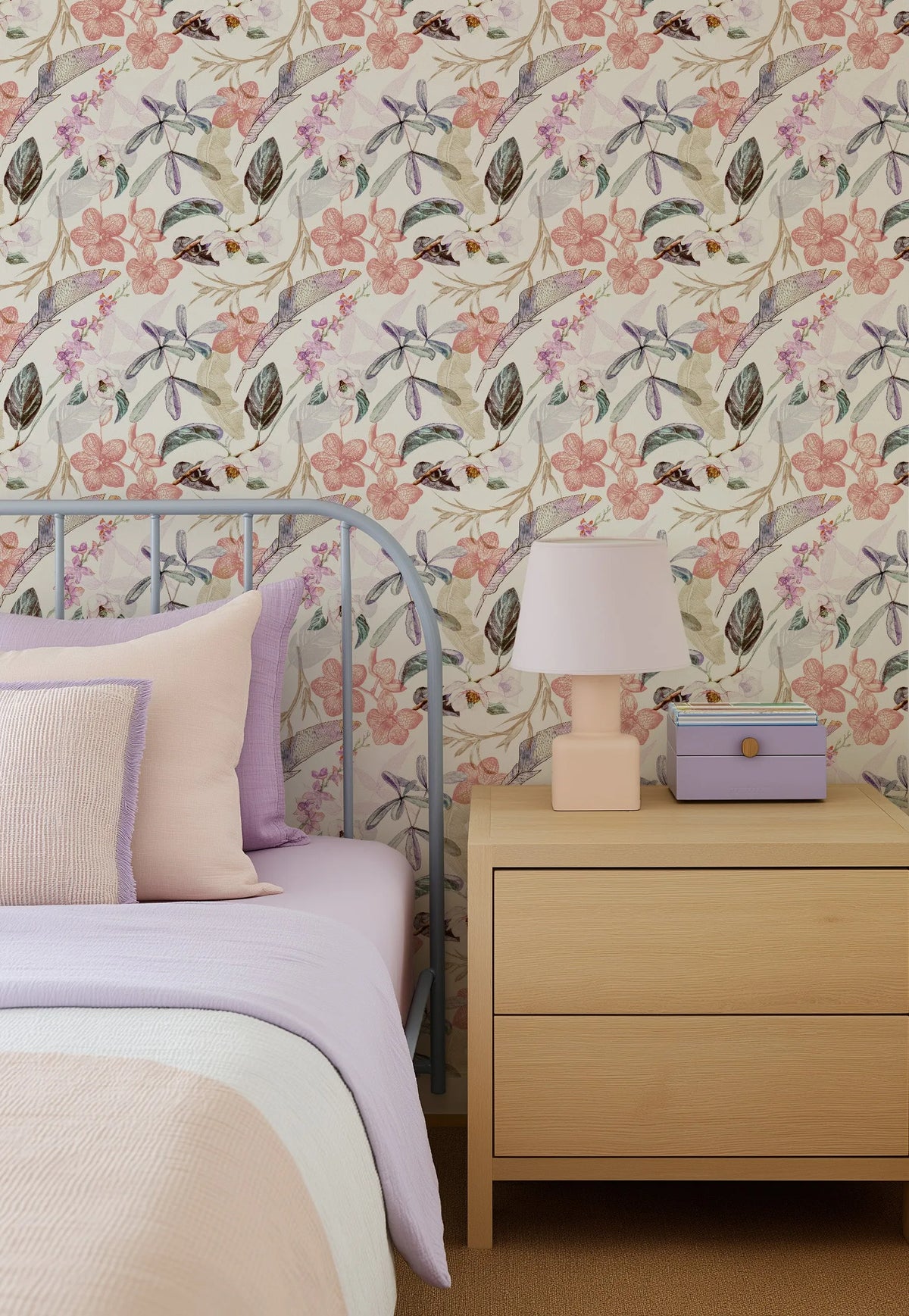 Oriental Orchids Wallpaper displayed on bedroom walls, featuring a repeating pattern of delicate pink and coral flowers, purple blossoms, dark green leaves, dragonflies, and small birds on a cream background. The wallpaper shows an illustrated botanical design with a soft, watercolor-like quality. A purple metal bed frame with lavender bedding and coordinating pillows is visible on the left, alongside a natural wood nightstand with a cream table lamp and purple decorative box.