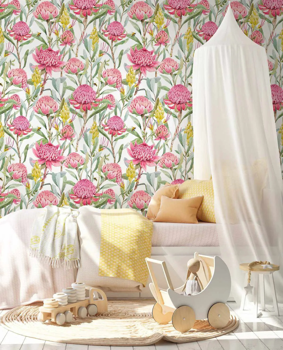 Lifestyle shot of a child's bedroom featuring Pink Waratah wallpaper. The wallpaper has a repeating pattern of hand-painted pink waratah flowers with green leaves and yellow flowering branches on a white background. The room includes a white bed with yellow and cream pillows and blankets, a white canopy, a light wood toy truck, and a white wooden toy pram with a stuffed bunny, all on a round woven rug.