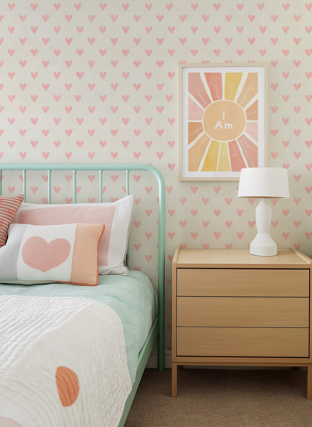 "Pink Hearts Wallpaper featuring a repeating pattern of small, hand-drawn blush pink hearts on a warm cream background, applied to the wall behind a mint green metal bed frame. The bed is dressed with a light green striped duvet, white textured quilt, and pillows in white, blush pink, and a white pillow with a large blush pink heart. A light wood nightstand with a white lamp sits beside the bed."