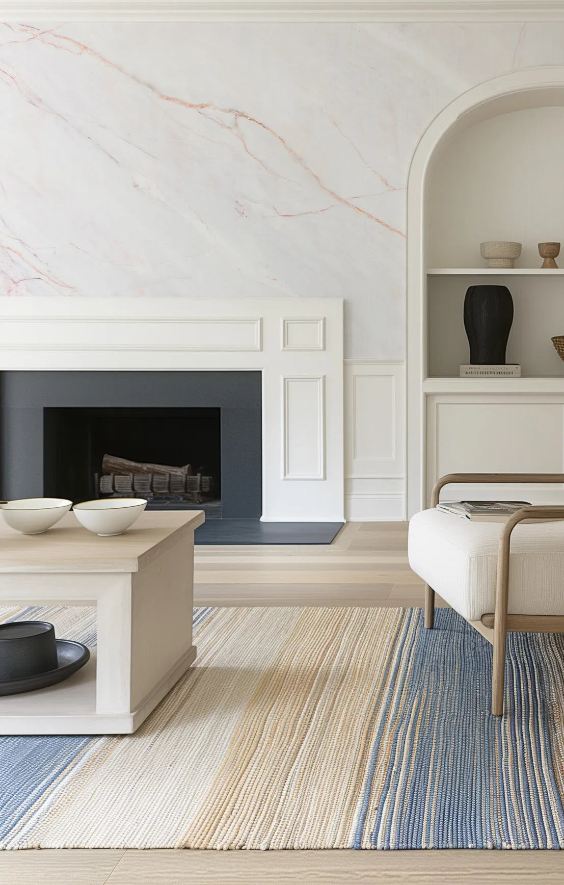 Lifestyle shot of a living room featuring peel and stick wallpaper with a white marble pattern and subtle rose gold veining above a white fireplace mantel. The room includes a light wood coffee table with two white bowls, a striped rug in shades of cream, beige, and blue, and a white upholstered bench with a light wood frame. A built-in arched shelf displays decorative objects.