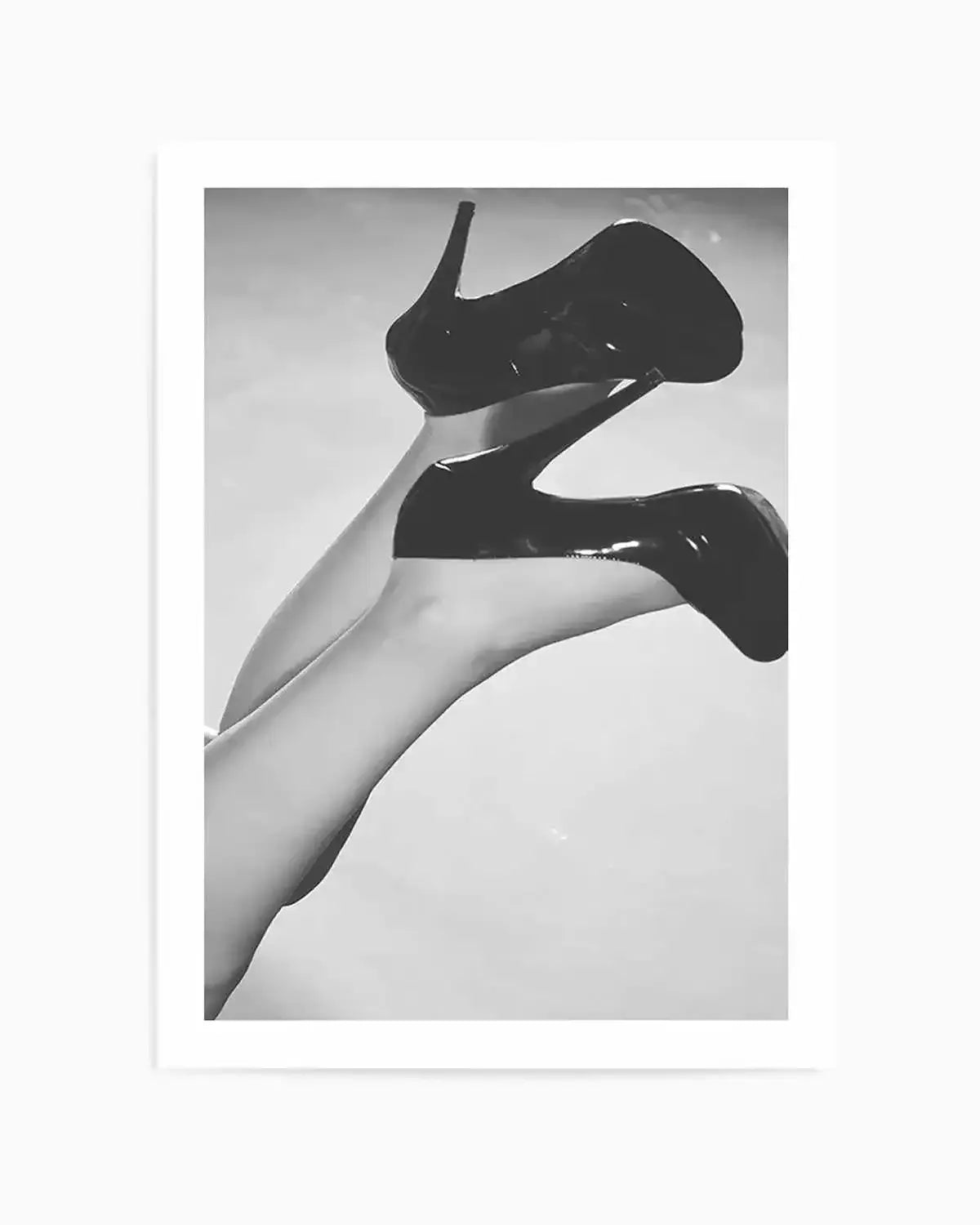 Framed black and white photograph of a person's legs and feet wearing high-heeled pumps, with the legs crossed and extended upwards against a light background. The glossy texture of the shoes is visible, contrasting with the smooth skin of the legs.