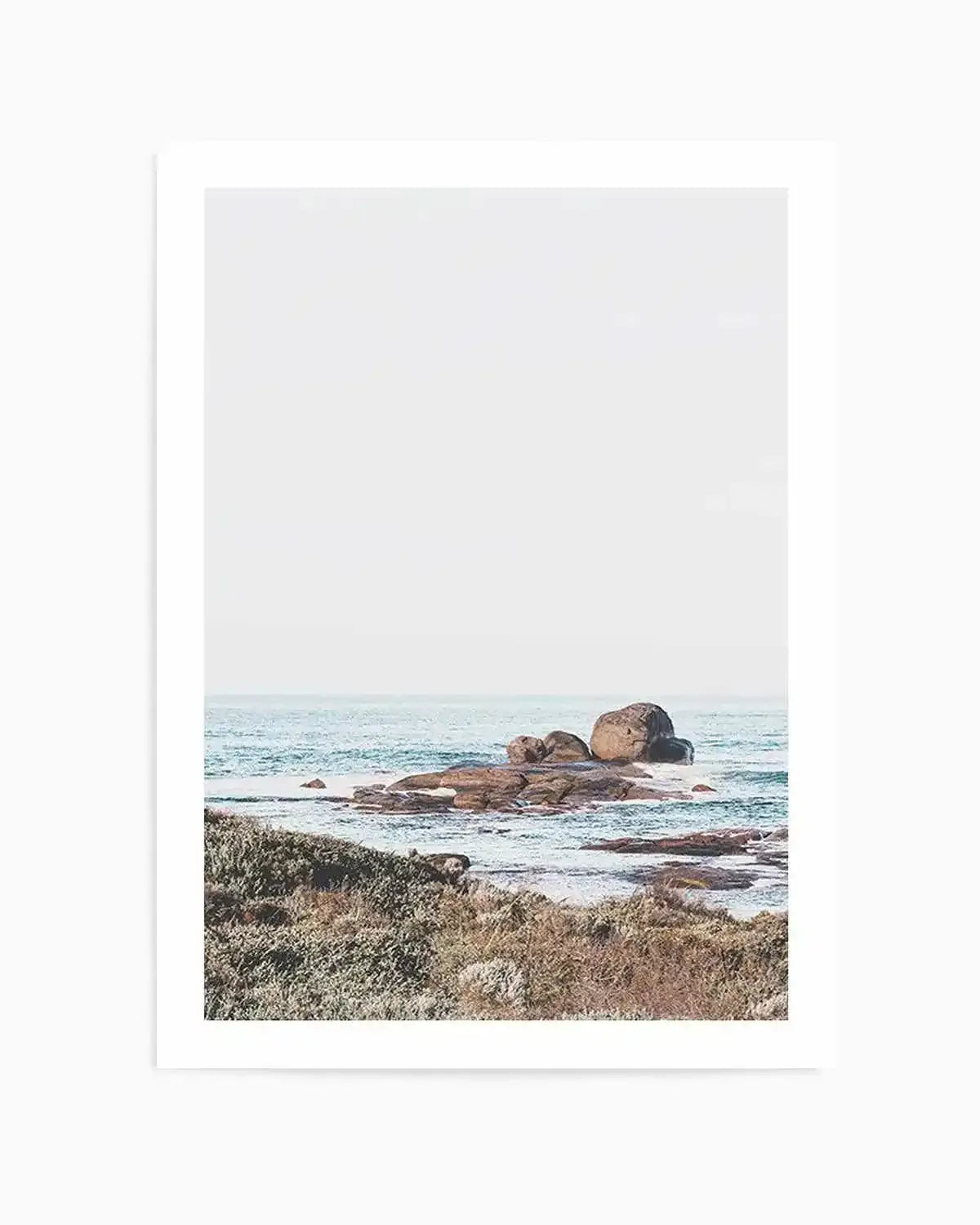 Framed photograph of a coastal scene featuring large, smooth boulders in the ocean with gentle waves and a light, hazy sky. The foreground shows dry, textured coastal vegetation in muted greens and browns. The print is displayed with a white border.