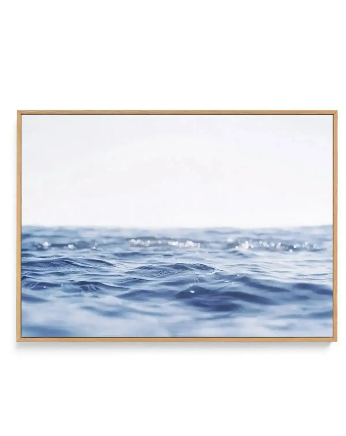 Framed canvas art print featuring a serene seascape photograph. The composition shows soft-focused ocean waves in shades of pale blue and deeper navy blue filling the lower two-thirds of the frame, with a misty white sky occupying the upper portion. The photograph has a calm, minimalist aesthetic with gentle wave textures and subtle light reflections on the water. The canvas is set in a natural wood-toned frame with a white border.