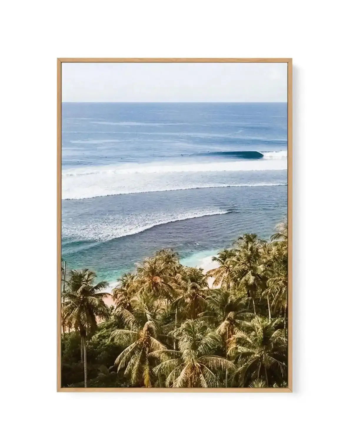 Framed canvas art print featuring a photograph of an Indonesian coastal landscape. The composition shows a foreground of lush green palm trees with detailed fronds, a middle section of turquoise shallow water and white sandy beach, and a background of rolling ocean waves in shades of blue with a surfer visible on a wave. Light gray sky occupies the top portion. The image is printed on canvas and set in a natural wood frame with a white border, creating a vertical landscape orientation.