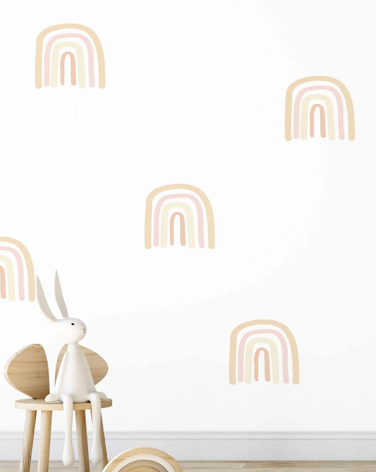 Lifestyle shot of a child's room featuring a white wall adorned with multiple simple bohemian rainbow decals in muted tones. The rainbows consist of five arches in pale yellow, blush pink, light beige, and soft peach. A white bunny toy sits on a wooden stool in the foreground, with a wooden rainbow toy visible on the floor.