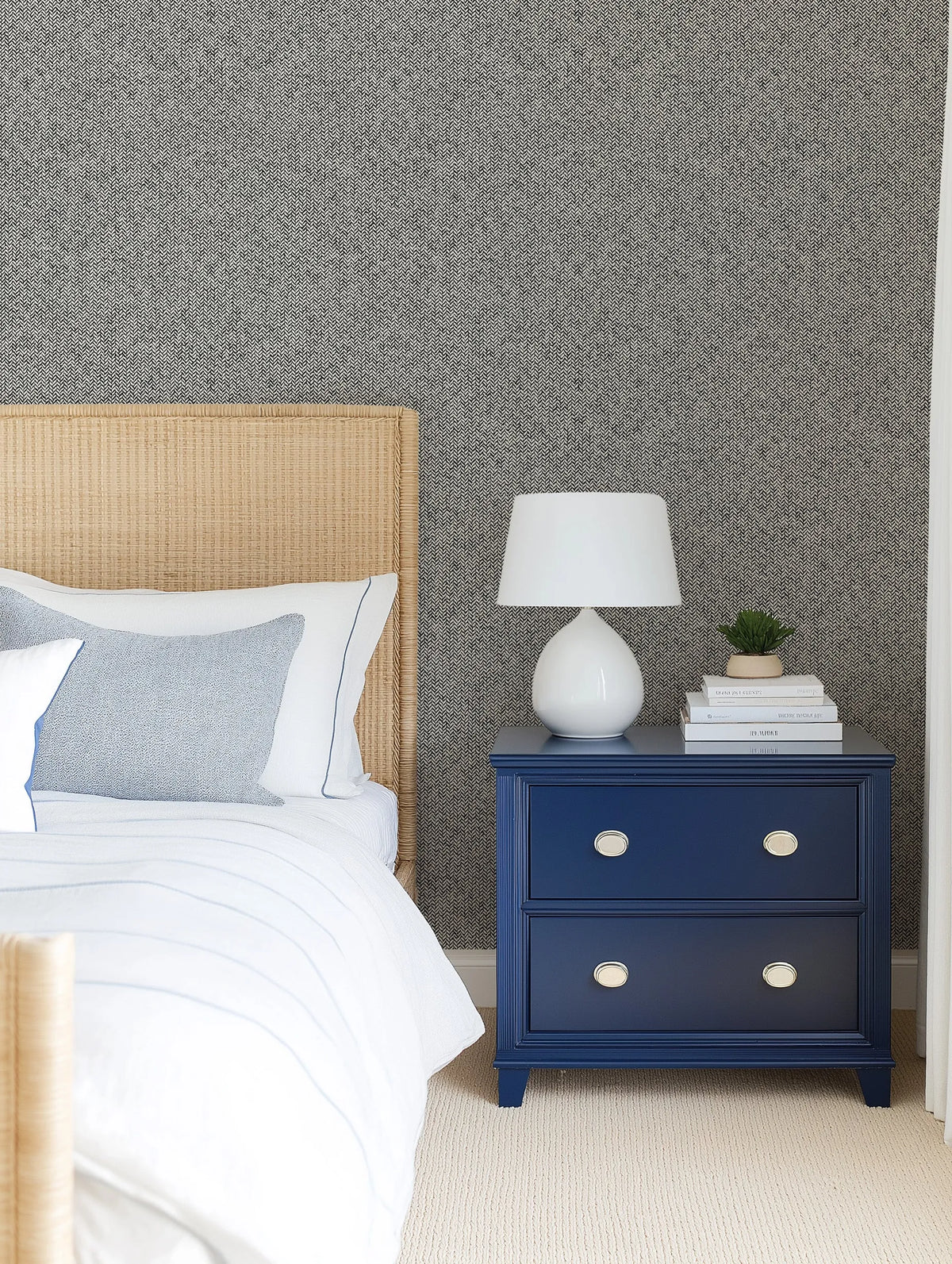Lifestyle shot of a bedroom featuring Soft Herringbone Wallpaper in a grey and off-white pattern. The wallpaper covers the wall behind a natural woven rattan headboard and a bed dressed in white and light blue striped bedding with a light blue textured pillow. A navy blue bedside table with silver oval knobs stands to the right, topped with a white ceramic lamp, a stack of white books, and a small potted succulent.