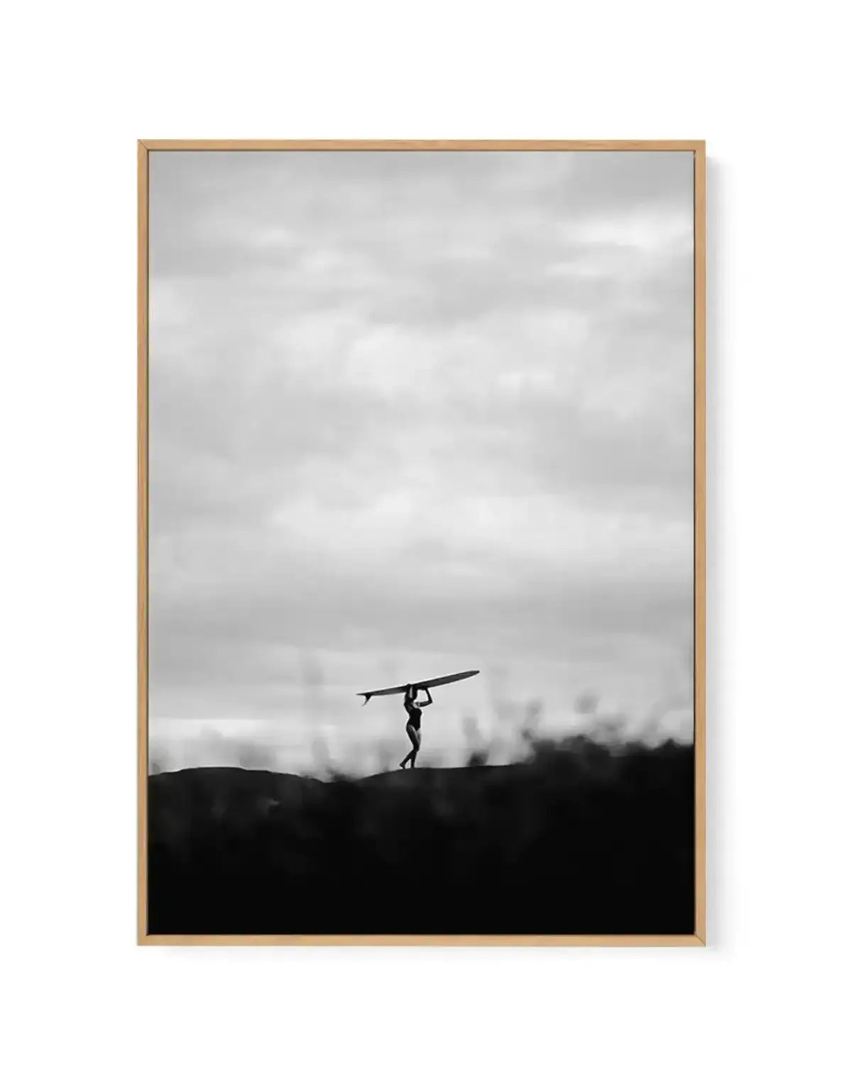Framed black and white canvas print of a lone surfer walking along a dark, silhouetted horizon against an overcast sky. The surfer, a small figure in the center, carries a surfboard over their head, with the dark foreground suggesting a dune or hill.