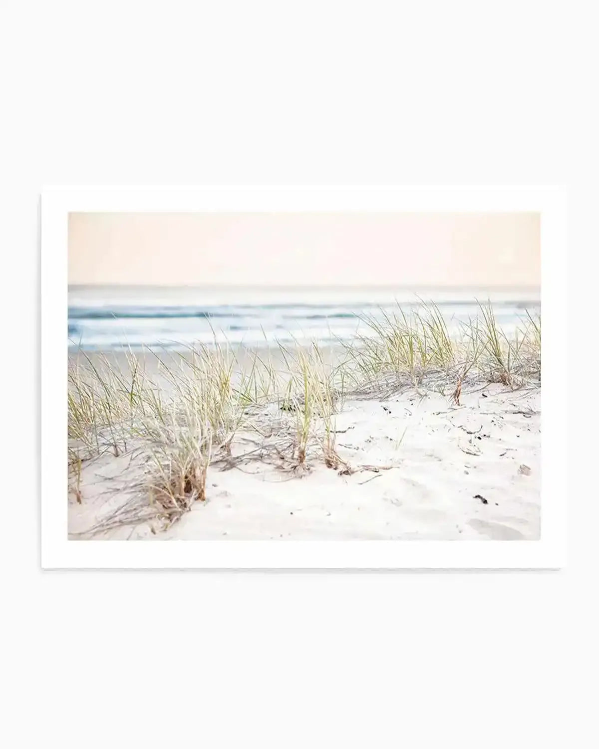 Framed photography art print of a serene beach scene with a soft, diffused light. The foreground features white sand dunes with sparse tufts of light green and golden beach grass. In the midground, gentle waves with white foam break on the shore, and the background shows a hazy, light blue ocean meeting a pale, almost white sky. The overall mood is peaceful and airy.