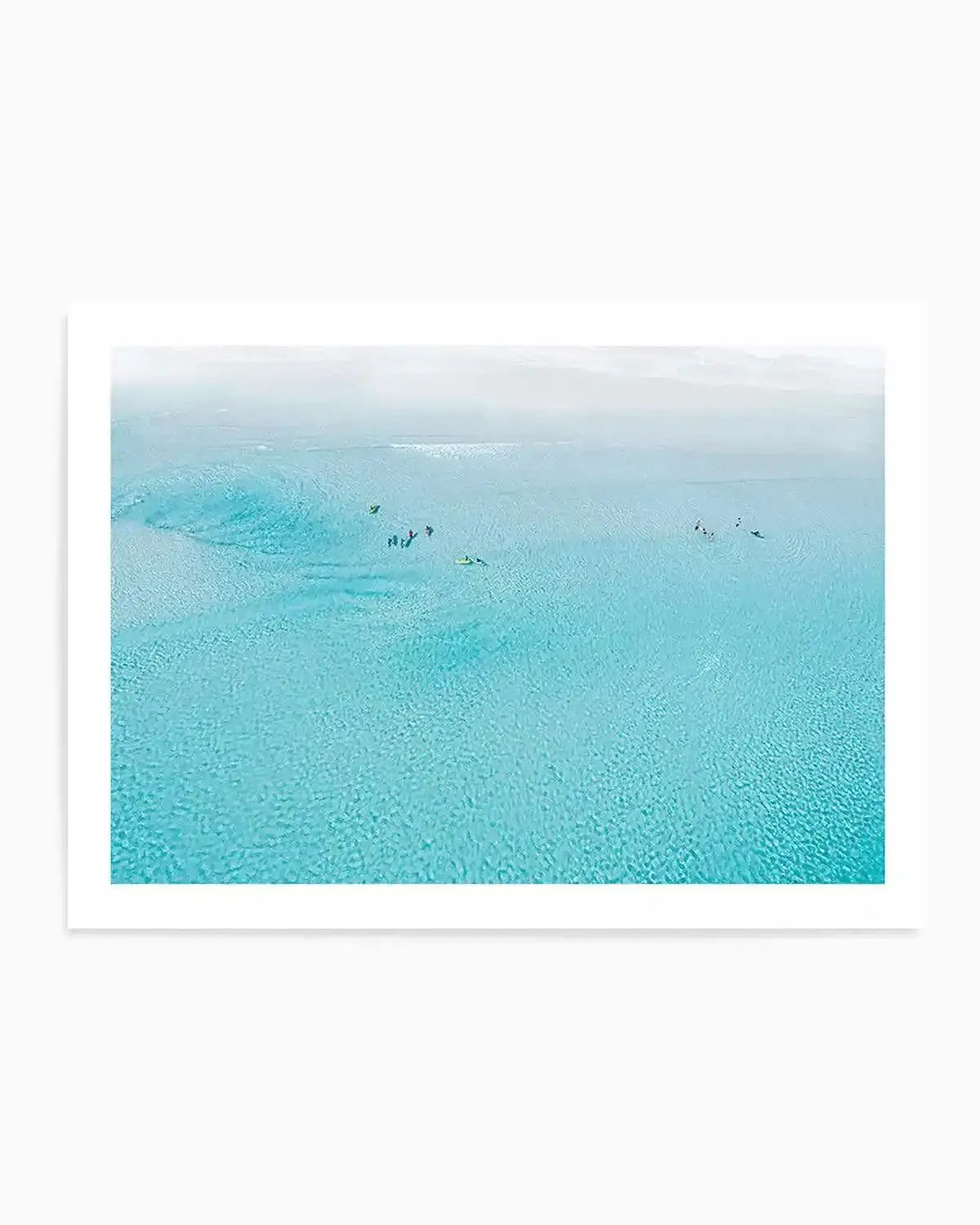 Aerial photography art print of a clear turquoise ocean with several small figures of people and surfboards scattered across the water, suggesting a surf school or group activity. The water shows subtle ripples and variations in depth, with a lighter, hazy horizon line. Printed on fine art paper with a matte finish.