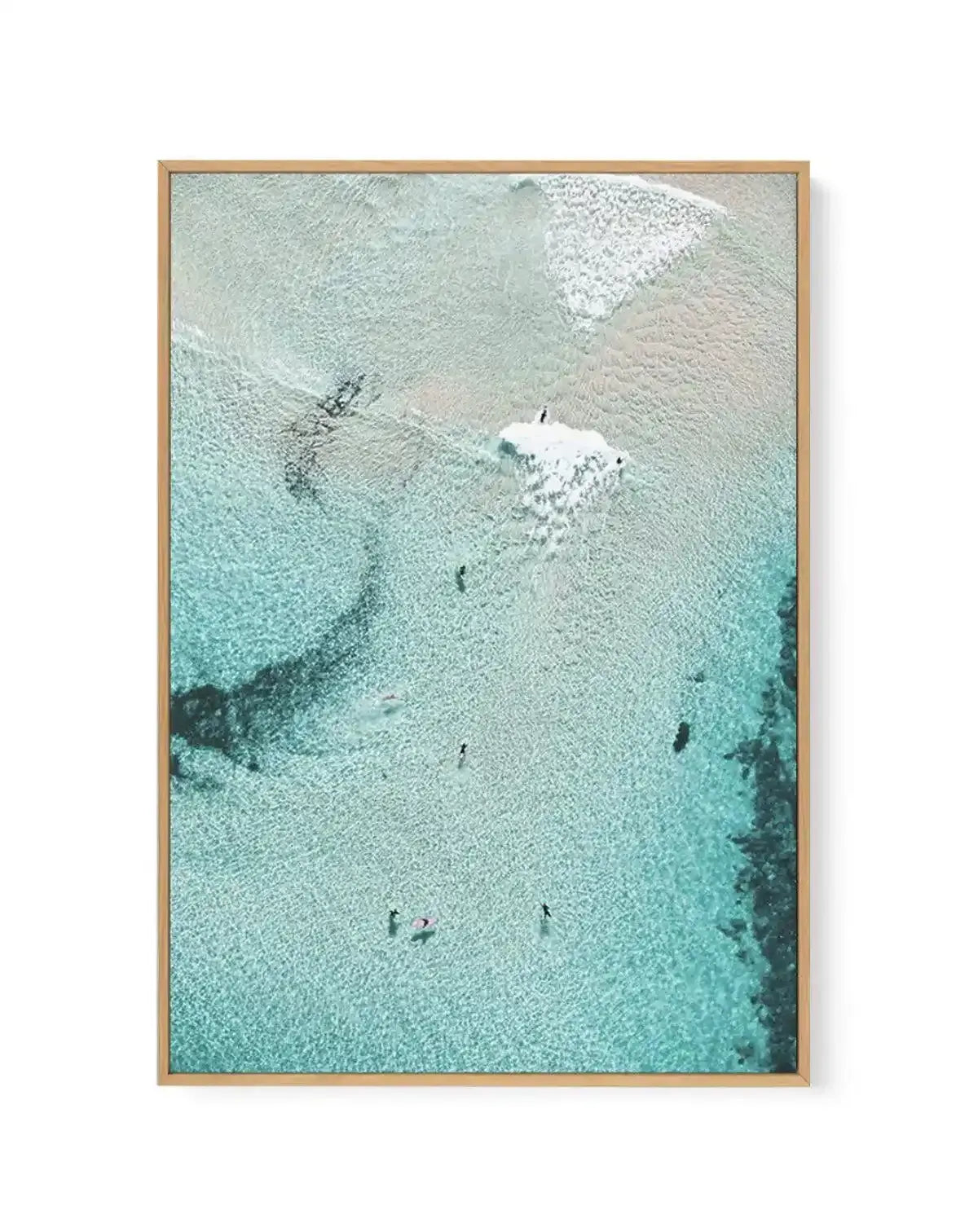 Aerial photograph art print of a clear turquoise ocean with gentle waves and several people swimming and surfing, displayed in a natural wood frame with a white border. The water reveals the sandy bottom and dark rock formations below.