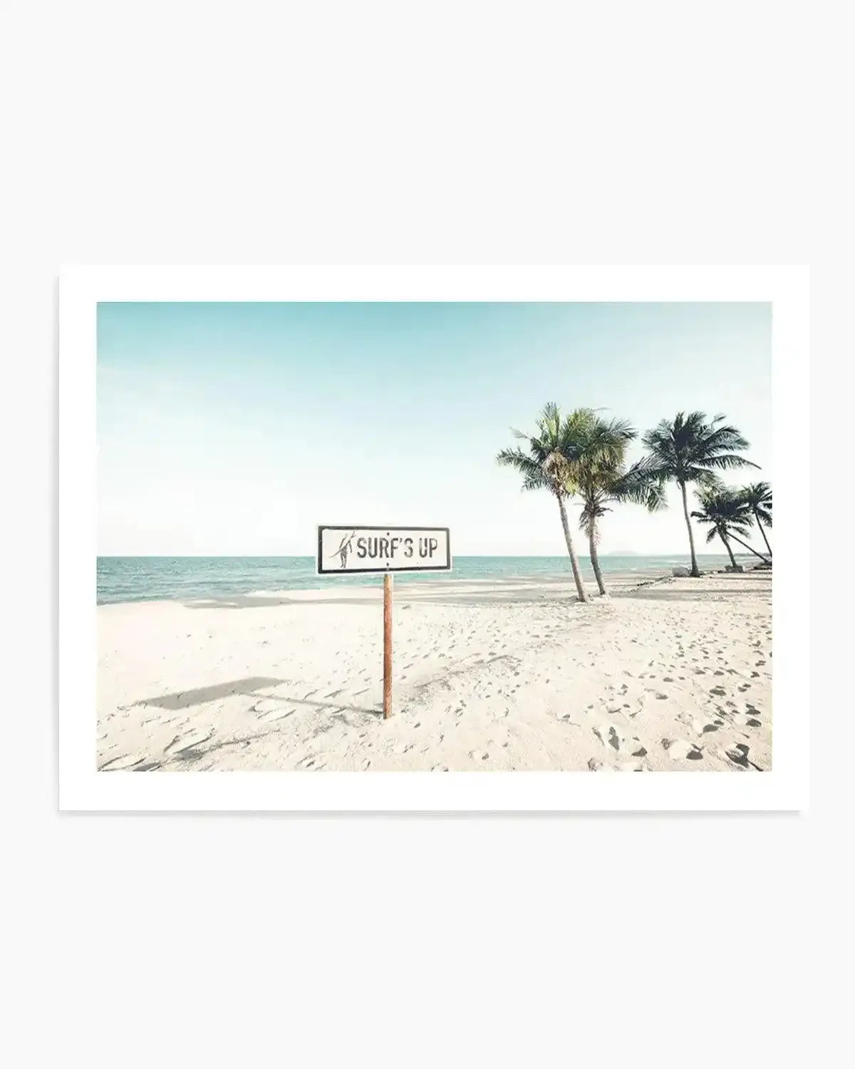 Framed photography art print of a serene tropical beach scene with a "SURF'S UP" sign, displayed in a white frame with a white border. The image features a wide expanse of light sandy beach with footprints, leading to calm turquoise ocean water under a clear pale blue sky. On the right, a cluster of palm trees with green fronds stands tall. A rustic wooden sign with a white rectangular face and black text reads "SURF'S UP" with a small black surfer icon, planted in the sand in the foreground.