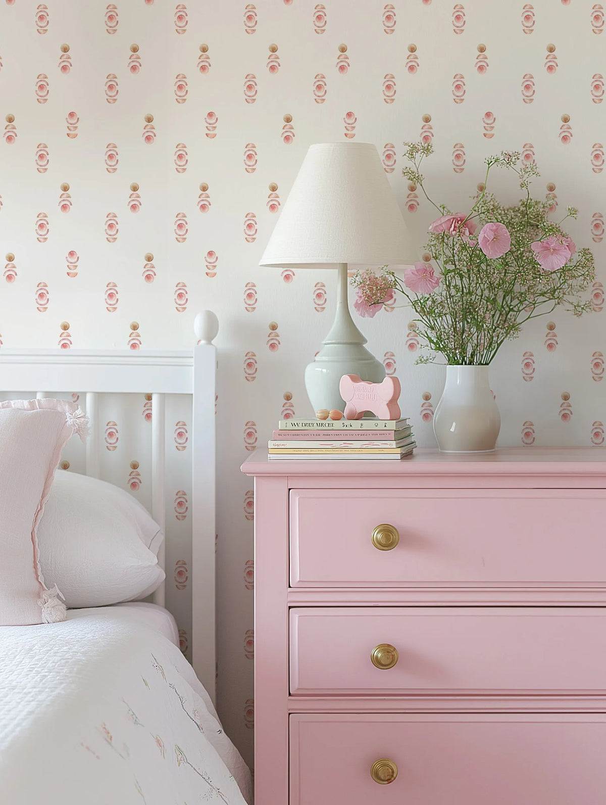 "Close-up of a white bed with pink pillows and a patterned quilt, next to a pink three-drawer dresser with gold knobs. On the dresser, a white lamp with a cream shade, a stack of books, a pink bone-shaped object, and a white vase filled with pink flowers and delicate greenery are displayed. The wall behind is covered in Sweet Boho wallpaper, featuring a repeating pattern of hand-painted abstract shapes in dusty rose and muted gold on a soft white background."