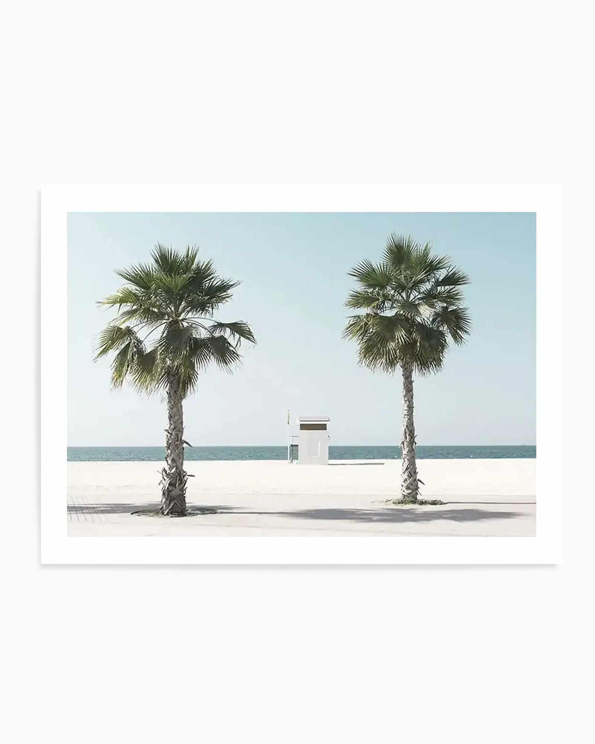 Art print depicting a minimalist beach scene with two tall palm trees flanking a small white lifeguard station on pale sand. The ocean and clear sky in soft turquoise tones create a serene, symmetric composition. The photograph-style artwork is presented in a white-bordered frame against a white background.