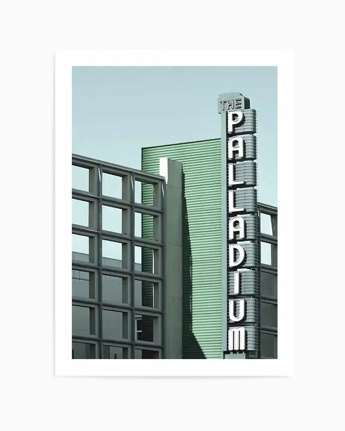The Palladium Art Print shows a vertical color photograph of a vintage neon sign reading "THE PALLADIUM" in white capital letters on a tall cylindrical metal structure. The sign is positioned against mid-century modern architecture featuring green corrugated metal siding and a grid-pattern window frame in dark gray. The sky is a pale blue-green. The photograph has a soft, desaturated color palette dominated by sage green and cool tones. The art print is presented with a white border on a white background.