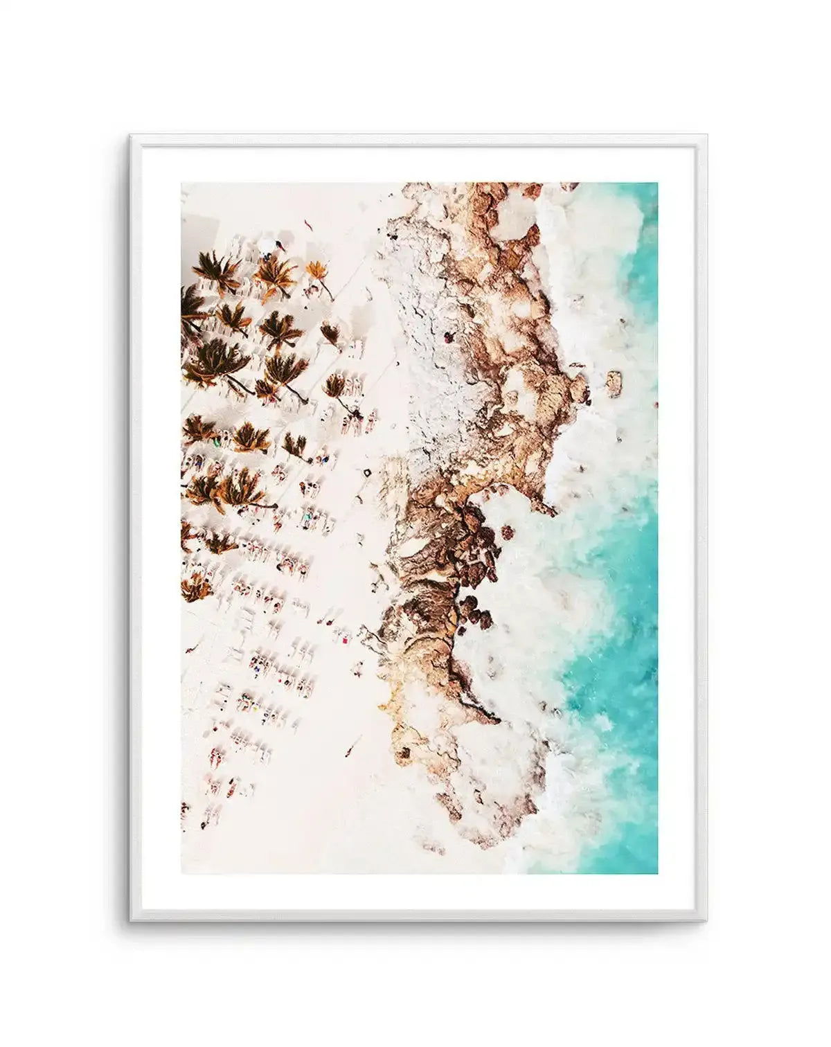 Framed aerial photograph of a tropical beach scene with a white frame and white border. The composition features a white sandy beach with scattered palm trees and tiny figures of people sunbathing and swimming, next to a rocky coastline, with clear turquoise ocean water on the right.