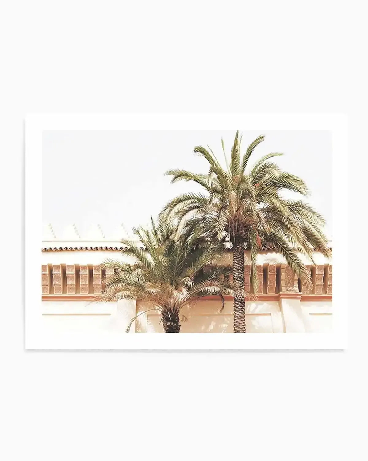 Photography art print featuring two palm trees in the foreground against a light-colored building with terracotta-colored details under a bright sky. The larger palm tree on the right has a tall, slender trunk and full fronds, while the smaller palm tree on the left is shorter and bushier. The building behind them has a decorative frieze with rectangular openings and a tiled roofline.
