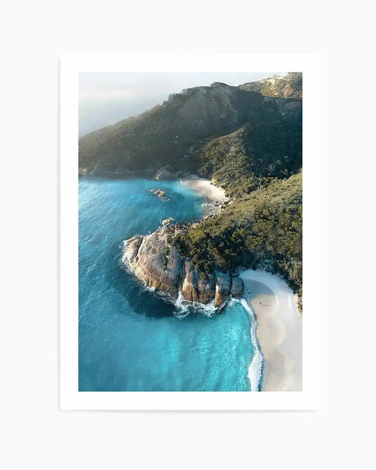 Aerial photography art print of a coastal bay with turquoise water, white sand beaches, and lush green hills. The composition features a large rocky outcrop dividing two crescent-shaped beaches, with gentle waves breaking on the shore and a hazy sky in the distance.