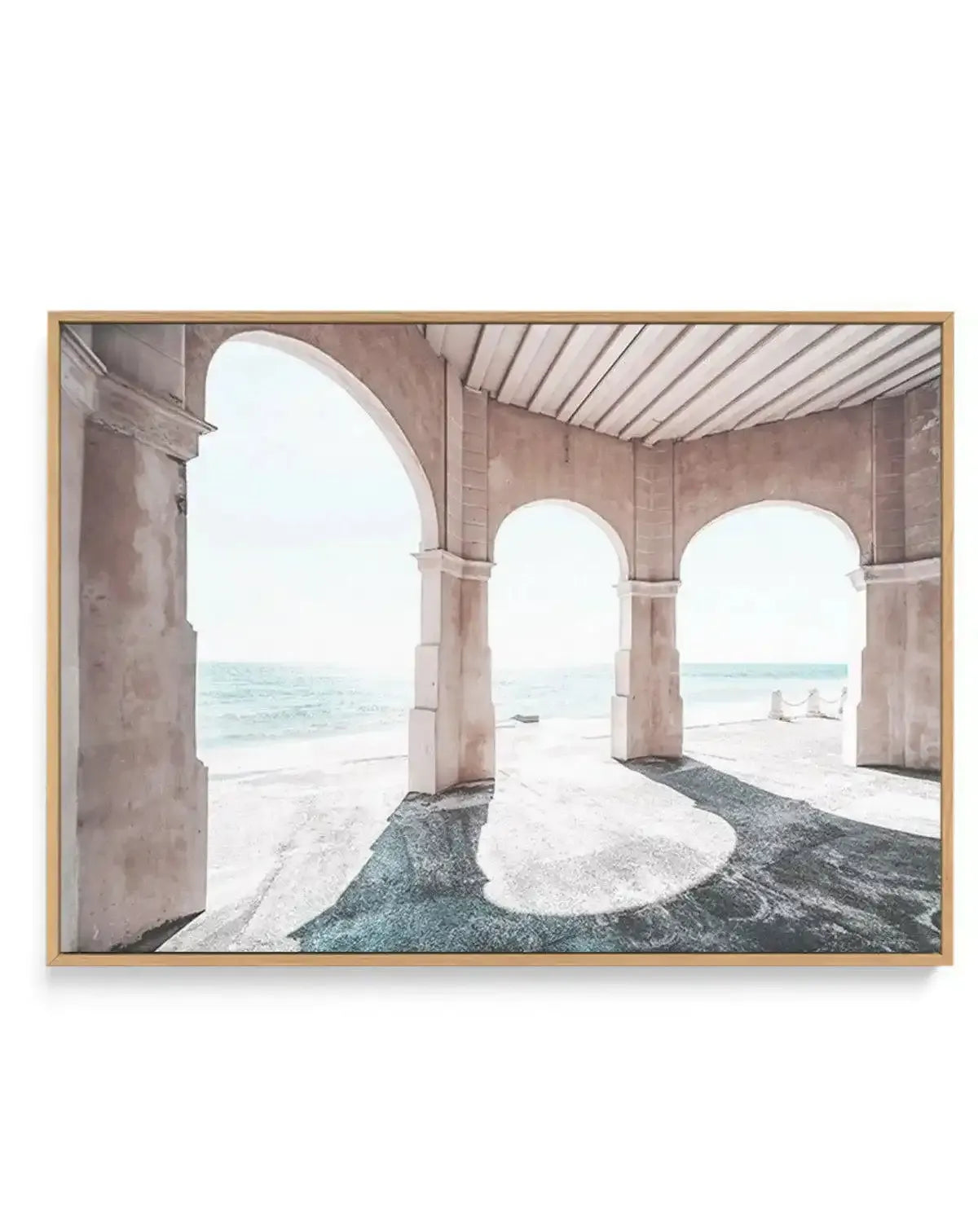 Framed canvas art print featuring a photograph taken from beneath a concrete pier at Cottesloe Beach. Three large arched concrete supports frame a view of calm turquoise ocean water and sandy beach beyond. The foreground shows wet sand in shades of teal and gray, while the sky is pale and overcast. The image has a serene, minimalist quality with soft natural lighting. The canvas is mounted in a natural wood frame with a light honey tone.