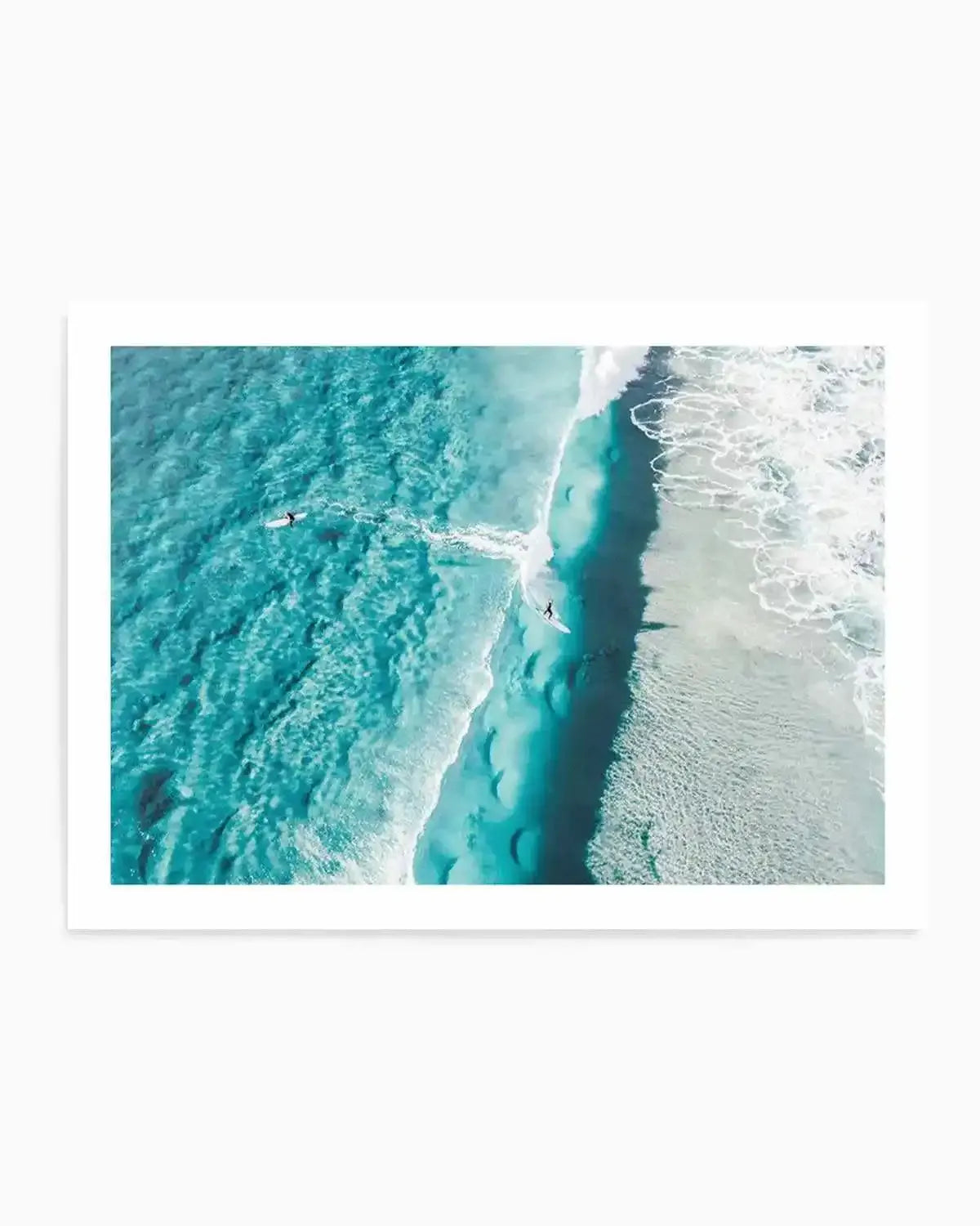 Aerial photograph art print of two surfers in turquoise ocean water near Mornington Victoria, with white foaming waves and a dramatic dark underwater trench visible from above. The image captures the contrast between shallow turquoise waters on the left, a deep navy channel in the center, and white-capped waves on the right shore. Printed on white paper with white border and displayed in a white frame.