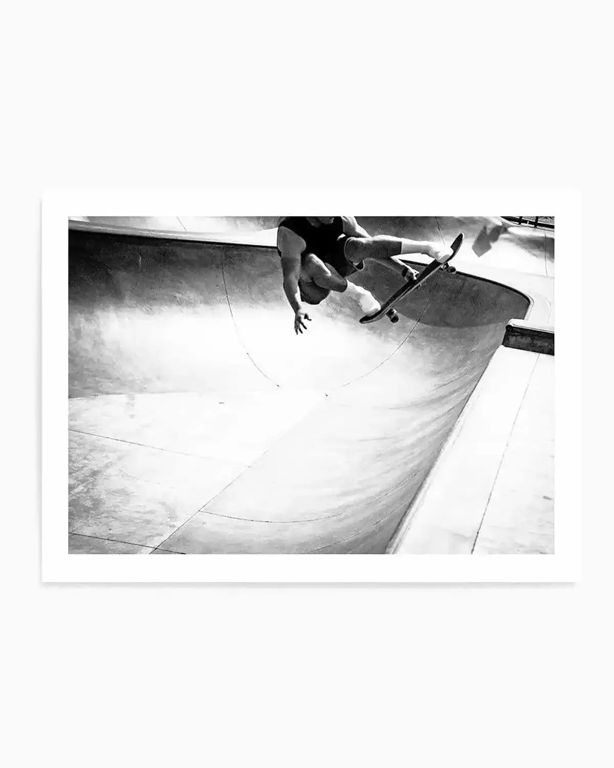 Framed black and white photograph of a skateboarder mid-air in a concrete skate park bowl. The composition captures the dynamic movement of the skateboarder, dressed in a dark top and shorts, with arms outstretched and the skateboard beneath their feet, against the smooth, curved surfaces of the park.