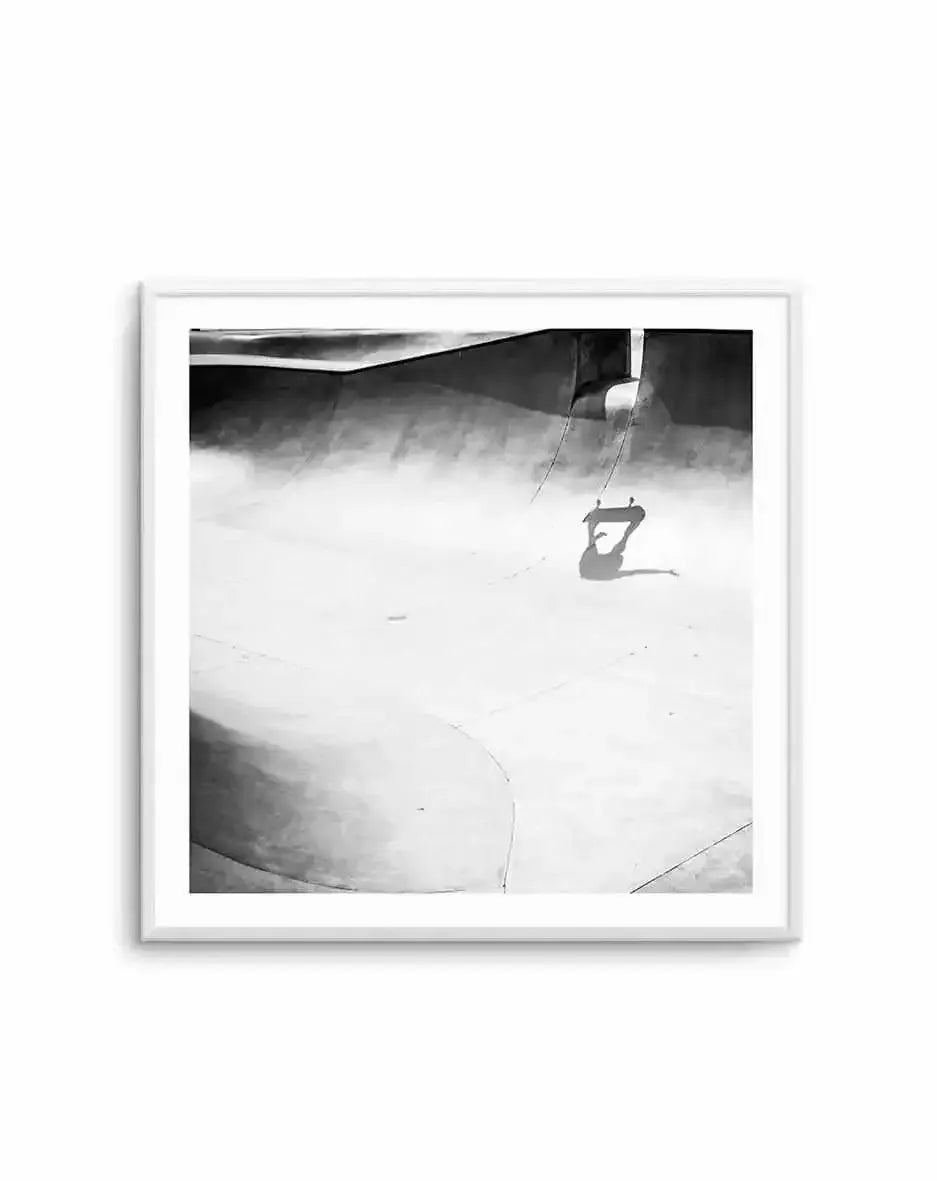 Black and white photograph framed in white showing a skateboarding figure suspended mid-air against a dramatic cloudy sky background. The skater appears as a silhouette performing a trick, hanging from ropes or cables attached to what appears to be a concrete or industrial structure above. The composition captures movement and contrast between the dark figure and bright, misty atmosphere. The print is displayed in a white square frame with white border.