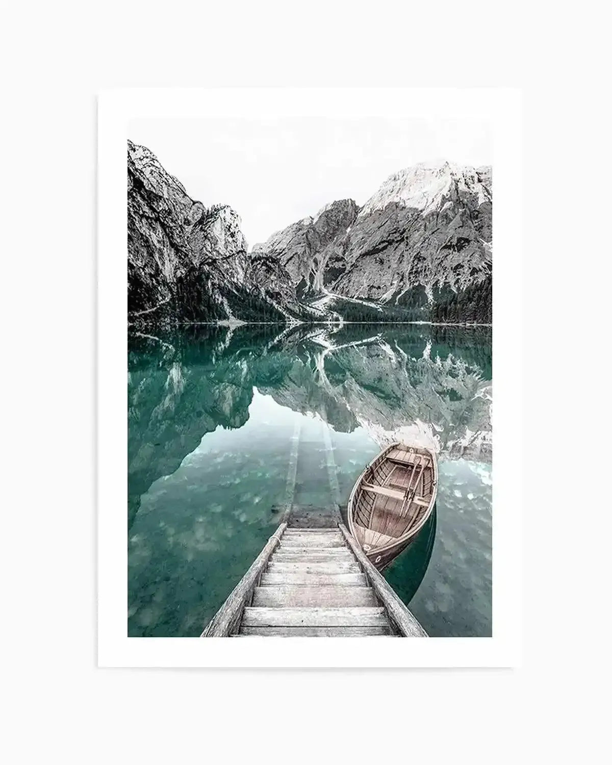 Framed black and white photograph of a mountain lake scene with a white border. The composition features towering, rugged mountains with snow-capped peaks reflecting in the calm, teal-hued water. A wooden boathouse dock extends into the foreground, with a classic wooden rowboat gently floating beside it.