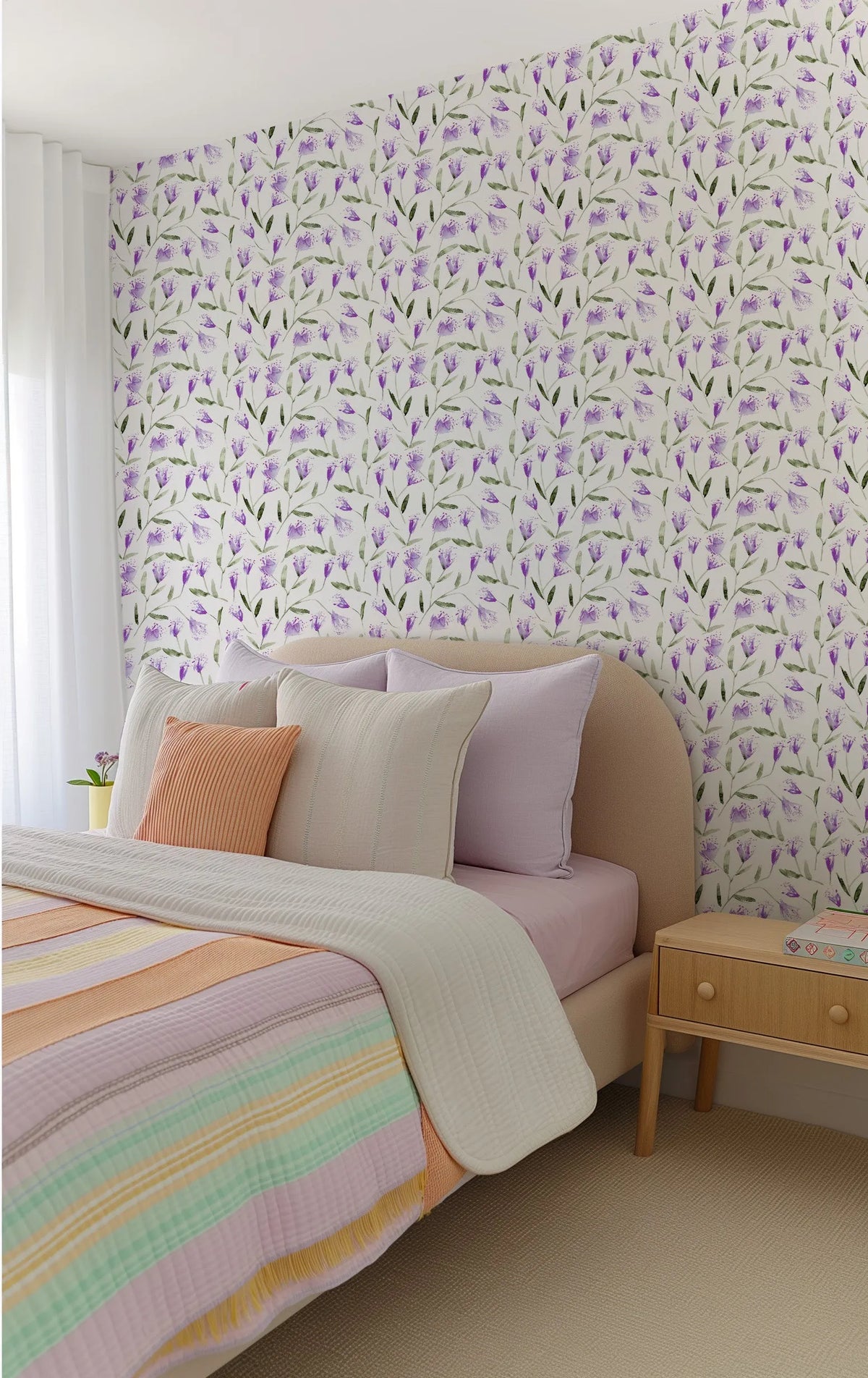 "A bedroom with a bed featuring a curved beige headboard, light pink sheets, and a striped quilt in pastel shades of pink, orange, yellow, and mint green. The wall behind the bed is covered in wallpaper featuring a repeating pattern of hand-painted violet flowers with green stems and leaves on a white background. A light wood nightstand with two drawers sits to the right of the bed, and a white curtain hangs on the left."
