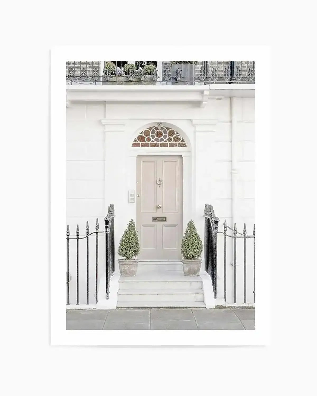 Framed photography art print of a white London townhouse entrance with a light beige door, natural wood frame, and white border. The door features a brass knocker and letterbox, with a decorative arched transom window above. Two small potted topiary trees flank the steps, and black wrought iron railings line the sidewalk.