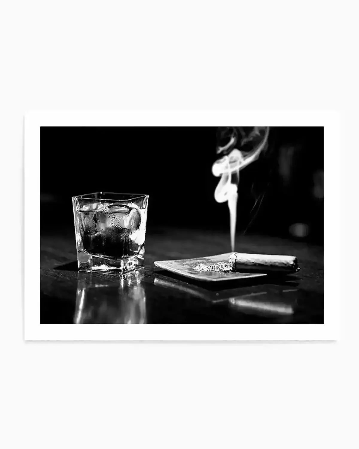 Black and white photograph showing a crystal tumbler filled with whisky and ice on the left, positioned next to a small plate holding a lit cigar with smoke rising upward. The composition is shot against a dark background with reflective surface, creating dramatic contrast and highlighting the textures of the glass and smoke. The art print is displayed with a white border frame.