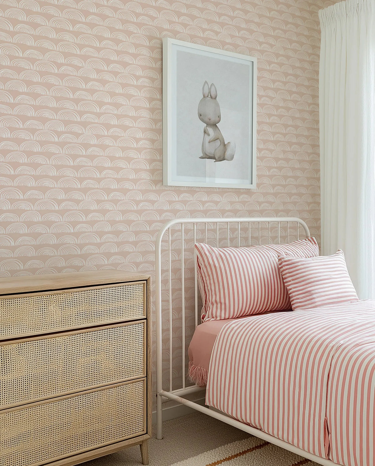Lifestyle shot of a child's bedroom featuring White Rainbows Wallpaper. The wallpaper has a repeating pattern of hand-drawn, arching rainbow shapes in off-white against a soft, dusty pink background. The wallpaper covers the wall behind a white metal bed frame with striped pink and white bedding, and a light wood dresser with woven cane drawers. A framed illustration of a grey bunny hangs above the bed.