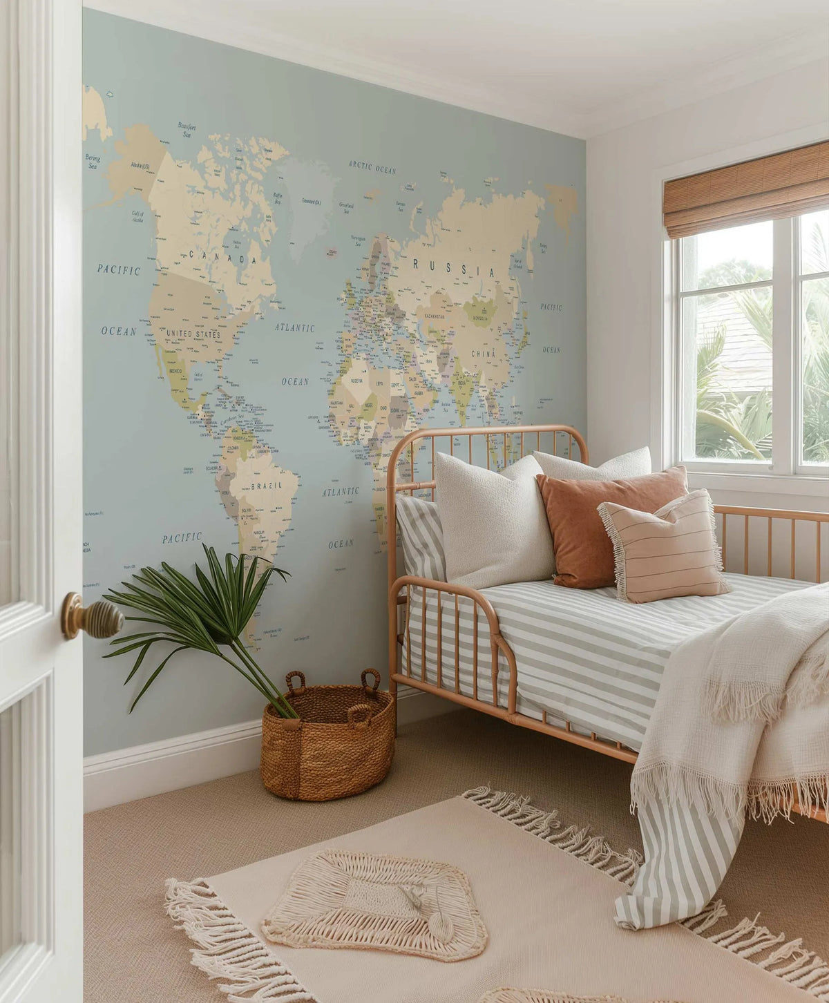 Peel and stick world map wallpaper mural covering an entire wall in a child's bedroom. The map features countries and oceans in muted tones of beige, light blue, and soft green, with country names and ocean labels in dark blue text. The room includes a light wood daybed with striped bedding, a textured cream throw, and various pillows in cream and terracotta. A woven basket with green palm fronds sits on the floor, which is covered in a light beige carpet with a cream rug.