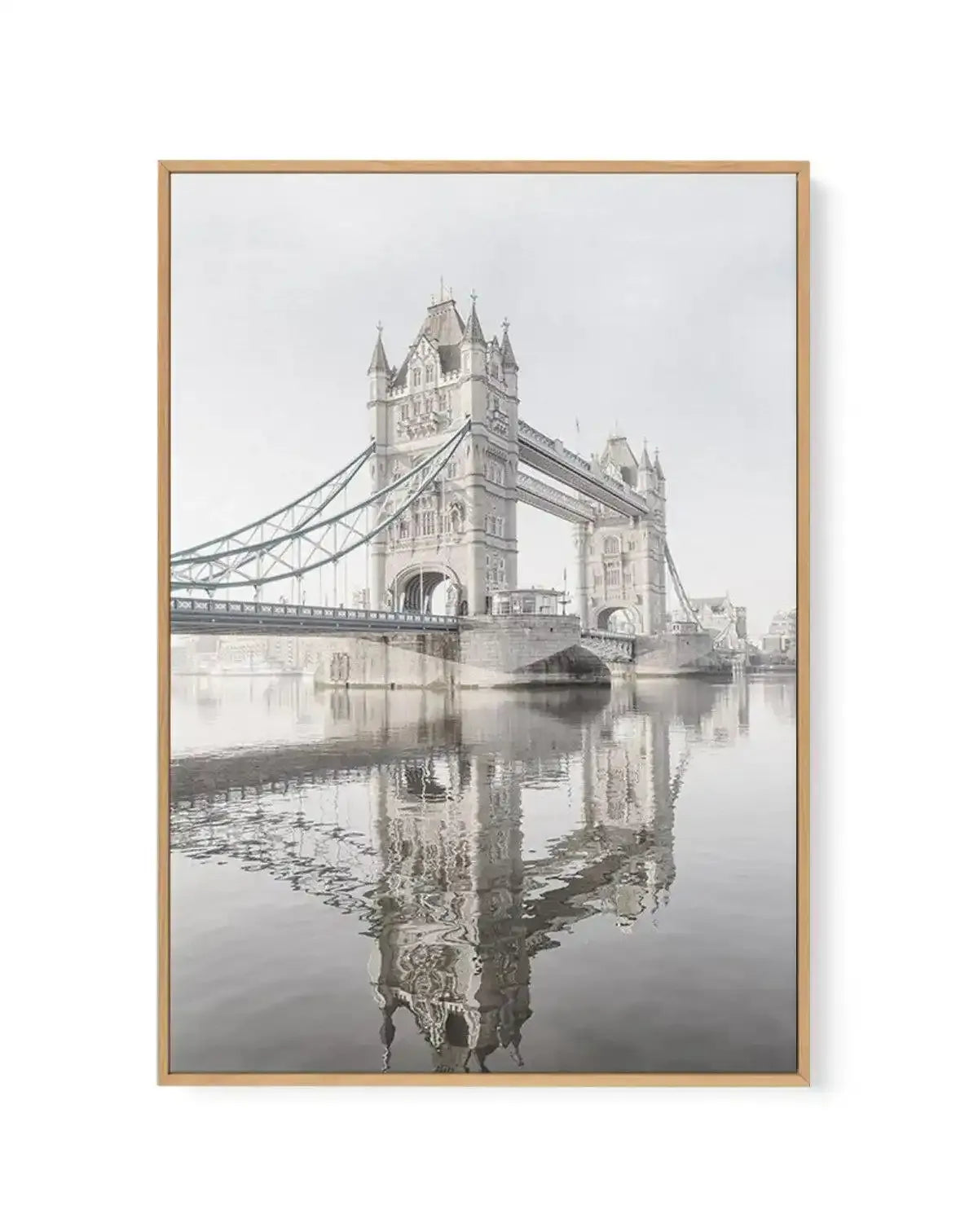 Framed black and white photograph of the Tower Bridge in London, printed on canvas and displayed in a natural oak frame. The composition features the iconic bridge with its two prominent towers and suspension cables, reflected in the calm water of the River Thames below, under a bright, overcast sky.