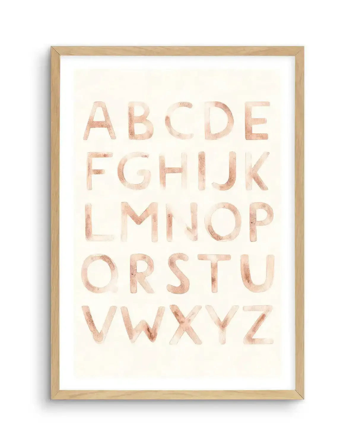 Watercolour art print featuring the complete alphabet in uppercase letters, painted in a soft, muted blush pink with visible brushstroke texture on a warm cream background, displayed in a natural oak frame with white border.