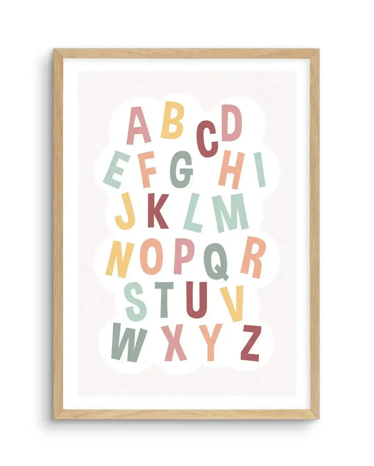 Alphabet art print featuring all 26 uppercase letters of the English alphabet in a playful, slightly irregular font, each in a different soft pastel colour, arranged in rows on a white background with subtle cloud shapes. The letters include shades of pale pink, soft yellow, muted teal, dusty rose, sage green, and light orange, displayed in a natural oak frame with white border.