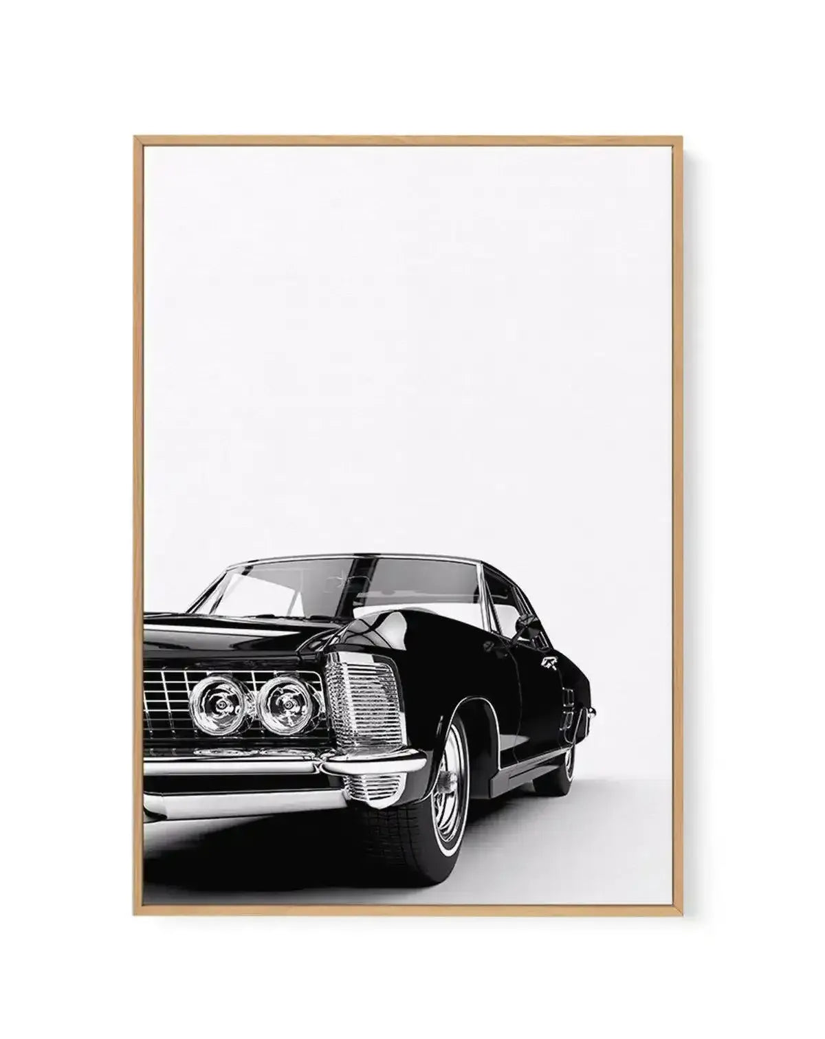 Black and white photograph of a classic American muscle car from the 1960s displayed in a natural wood frame. The car, positioned at a three-quarter angle facing left, features dual round headlights, a prominent vertical grille, and a sleek body with the driver's door open. The image has a minimalist composition with ample white space above the vehicle against a clean white background. The wood frame has a warm tan finish with a simple, modern design.