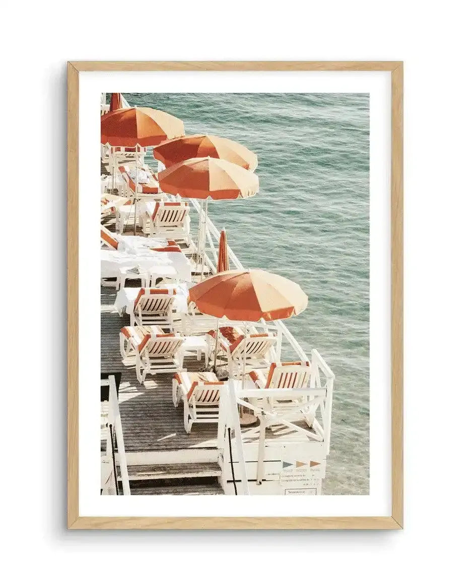 Framed art print featuring a vintage photograph from 1972 showing a ship's deck lined with white lounge chairs and orange-and-cream striped umbrellas overlooking turquoise-green ocean water. The photograph has warm, saturated colors with a nostalgic quality. Displayed in a natural wood frame with white border, evoking mid-century leisure and travel aesthetics.