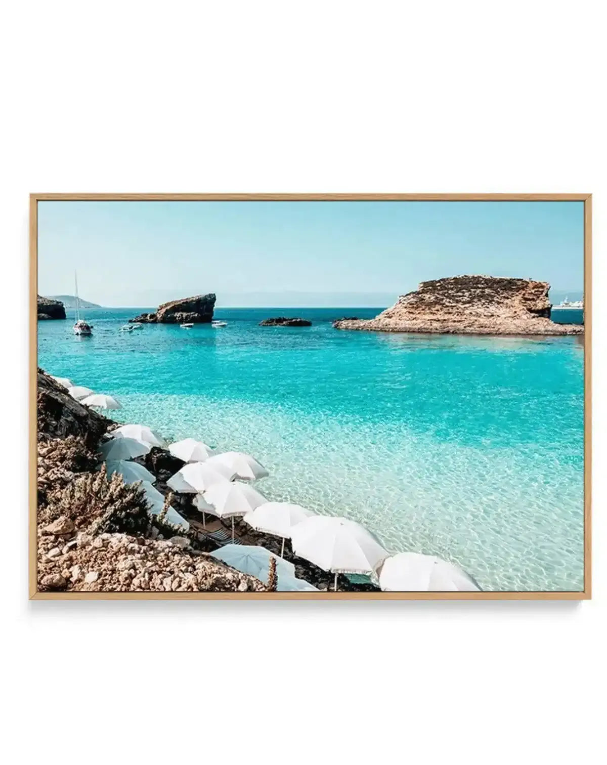 Framed horizontal canvas print featuring a Mediterranean beach scene in Blue Lagoon, Malta. The photograph shows crystal-clear turquoise water with rocky outcrops and small islands, a row of white beach umbrellas along the foreground rocks, and sailboats anchored in the shallow bay. The sky is pale blue and cloudless. The canvas is mounted in a natural wood frame with a light tan finish.