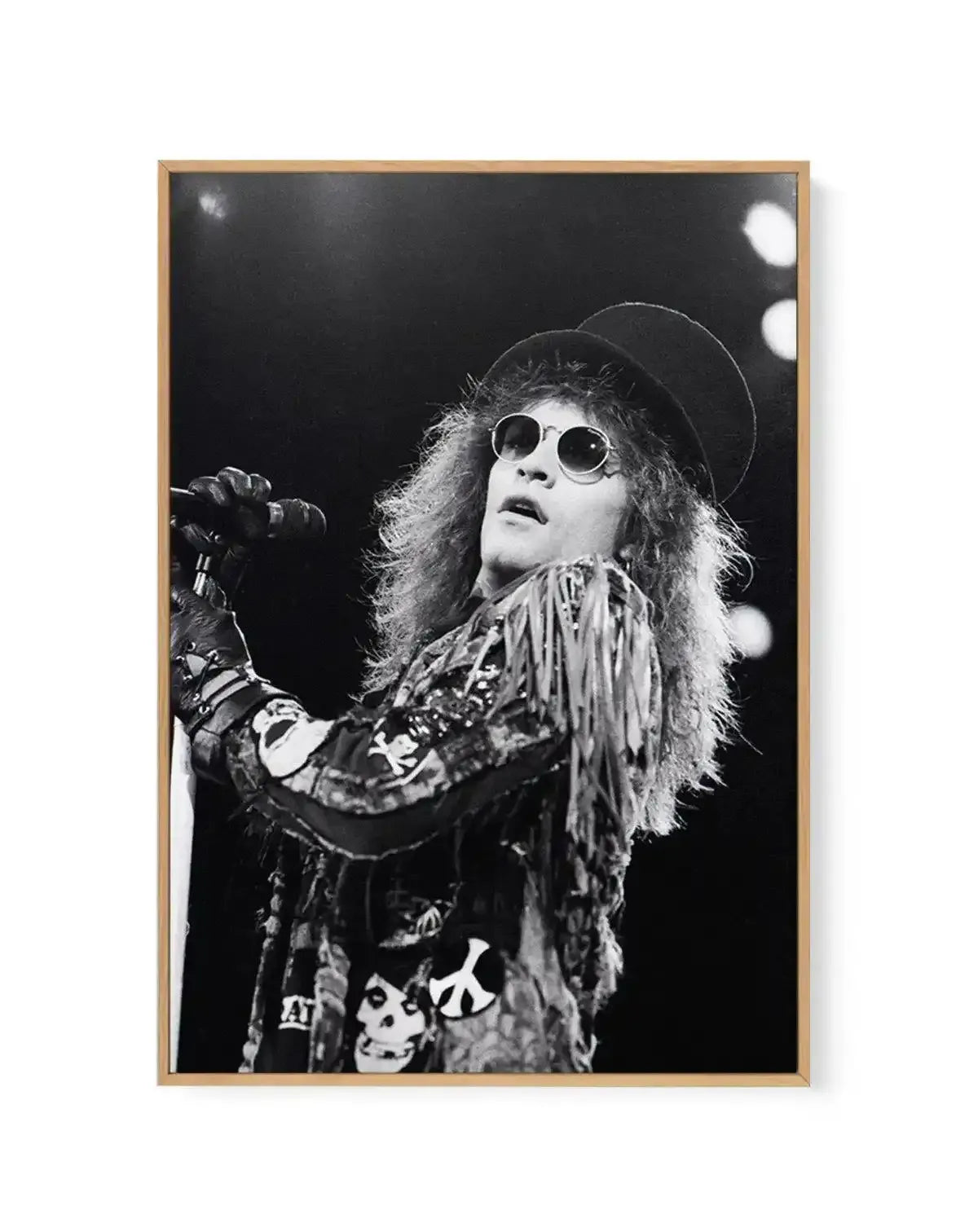 Framed black and white photograph of Bon Jovi frontman Jon Bon Jovi performing on stage, printed on canvas and displayed in a natural wood frame. The composition captures Bon Jovi mid-song, looking upwards with a microphone in his gloved hand. He wears a hat, round sunglasses, and a fringed jacket adorned with patches including a peace sign and skulls. The background is dark with blurred stage lights.
