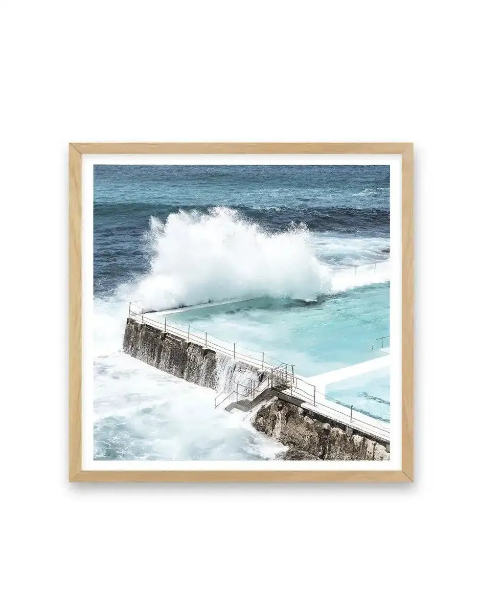 Framed photograph of the iconic Bondi Icebergs pool in Sydney, Australia, with a large wave crashing over the edge into the turquoise pool water. The natural oak frame has a white border, and the image captures the dynamic movement of the ocean against the concrete pool structure.