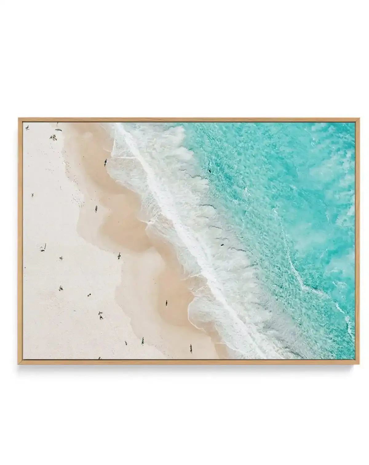 Aerial photograph of a sandy beach meeting turquoise ocean water, with white foaming waves and scattered swimmers visible on the pale sand. The image transitions from cream and beige tones on the left beach area to vibrant aqua and teal water on the right. Printed on canvas in a natural wood frame with a horizontal landscape orientation.