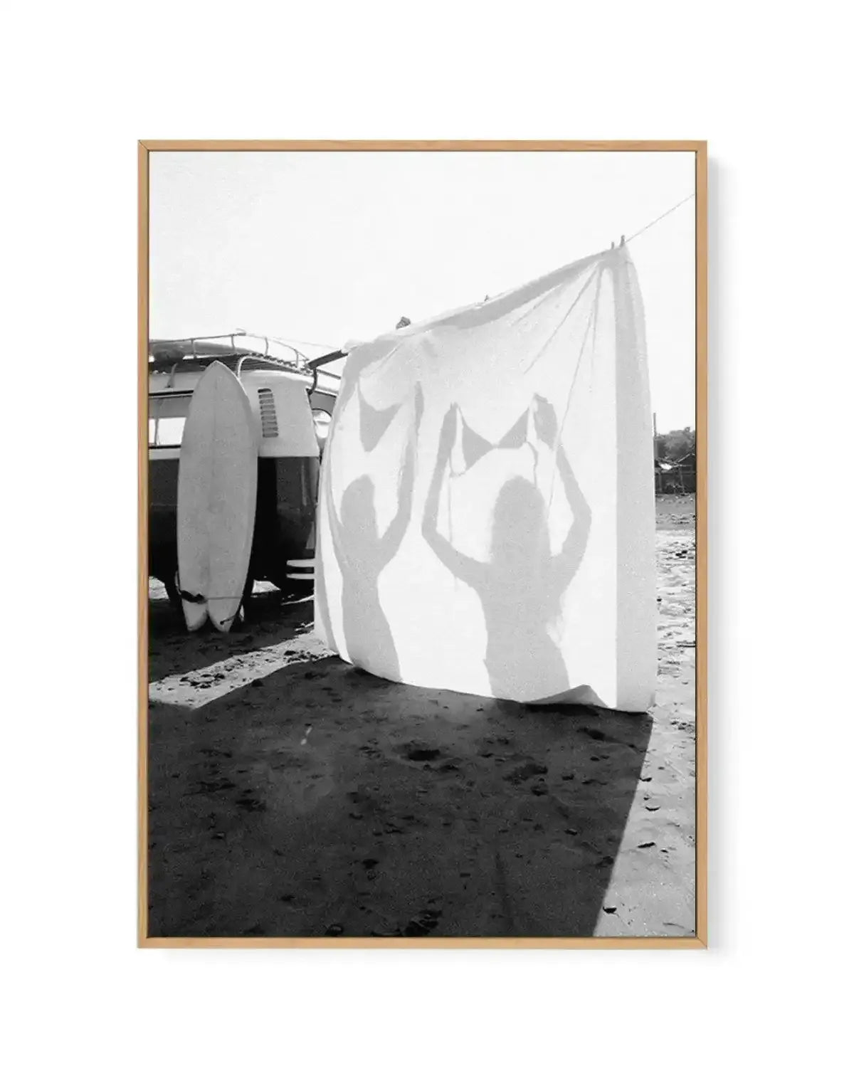Black and white framed canvas art print titled "Born Free" by Mario Stefanelli, displayed in a natural wood frame. The photograph shows a white fabric or sail billowing in the wind on a sandy beach, with silhouettes of two figures visible through the translucent material. A vintage vehicle and surfboard are positioned to the left. The composition captures movement and freedom against a bright sky, printed on canvas with a white border, mounted in a warm tan wooden frame.
