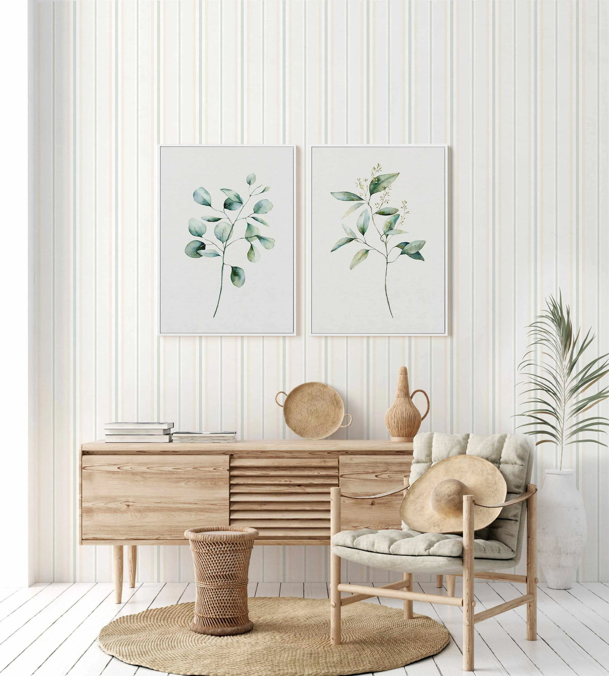 "A living room featuring Byron Stripe Wallpaper with thin vertical stripes in pale sage green, light beige, and off-white. Two framed botanical art prints hang above a light wood console table. The room includes a woven straw rug, a wicker basket, a light wood chair with a cream cushion and straw hat, and a tall white vase with a large green plant."