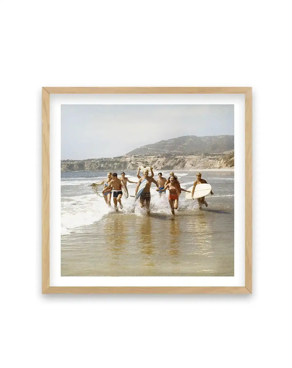 Framed art print featuring a vintage color photograph of a group of young people running into the ocean at a sandy beach, some carrying surfboards, with coastal cliffs and hillside visible in the background under a hazy sky. The photograph has warm, muted tones with golden and soft blue hues. The print is displayed in a natural wood frame with a white border.