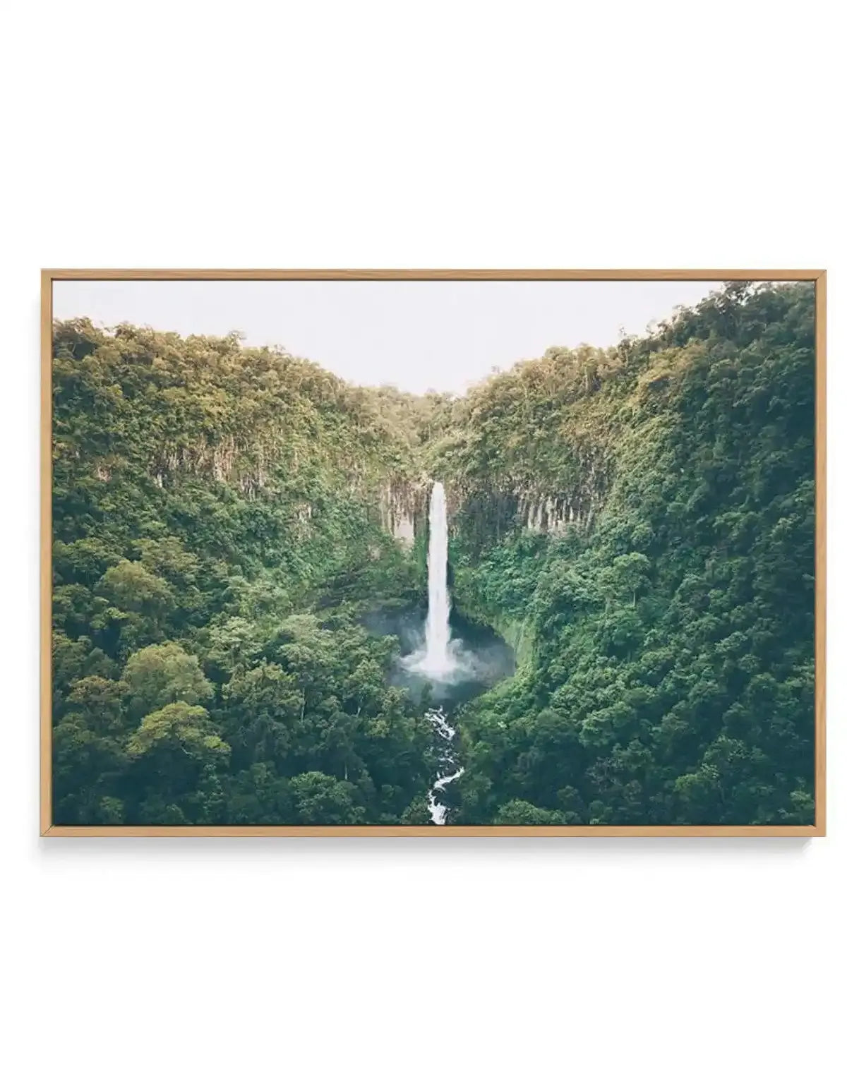 Framed canvas art print of Cannabullen Waterfall featuring a tall, slender waterfall cascading down a lush green valley surrounded by dense forest vegetation. The composition shows the waterfall in the center with steep, forested cliff walls on either side covered in varying shades of green. A white stream flows down the rocky base below the falls. The sky above is overcast and pale. The canvas is mounted in a natural wood frame with a light tan finish.