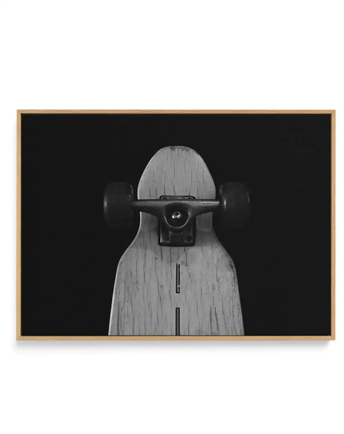 Black and white photograph of a wooden skateboard deck shown front-on against a dark background. The deck features natural wood grain texture with visible vertical striping down the center. A black skateboard truck with metal hardware is mounted horizontally across the upper portion. The image is printed on canvas and displayed in a natural wood frame with a light tan finish.