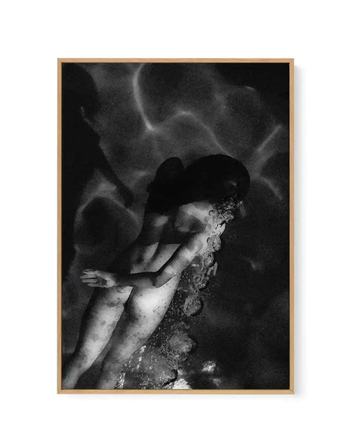 Framed black and white photograph of a person submerged underwater, with a natural wood frame and white border. The composition shows the person's body from the waist up, with their back to the viewer and arms crossed in front. Bubbles rise from their head, and streaks of light ripple across the dark water, creating a mysterious and serene mood.