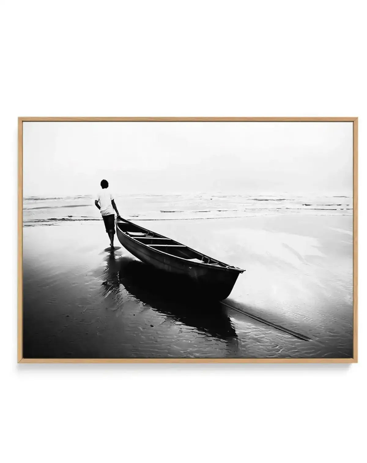 Black and white photograph in natural wood frame showing a shirtless fisherman standing beside a dark wooden boat on a wet beach. The man holds the boat's rope while facing the calm ocean horizon. Wet sand reflects the figure and vessel. Overcast sky creates a muted, contemplative atmosphere. Fine art photography print with subtle gradient tones from dark blacks to light grays. Natural wood frame with white border.