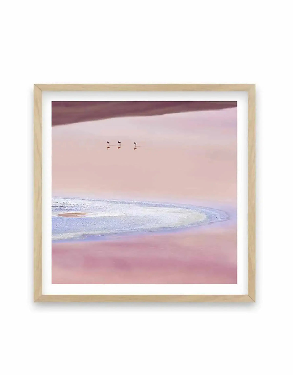 Framed art print featuring a minimalist landscape photograph of a pink lake with three pairs of flamingos in the distance, displayed in a natural oak frame with white border. The lake features a curved white and blue shoreline, with the water transitioning from light pink to a deeper magenta.