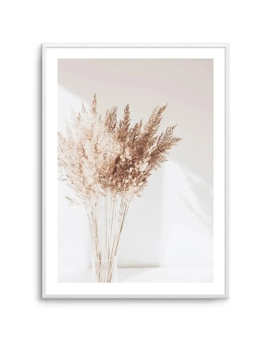 Framed photography art print featuring a bouquet of dried pampas grass in a clear glass vase, with a white frame and white border. The pampas grass has feathery plumes in shades of light beige and warm brown, with long, thin stems. The background is a soft, light cream color with subtle shadows and highlights, creating a serene and minimalist mood.