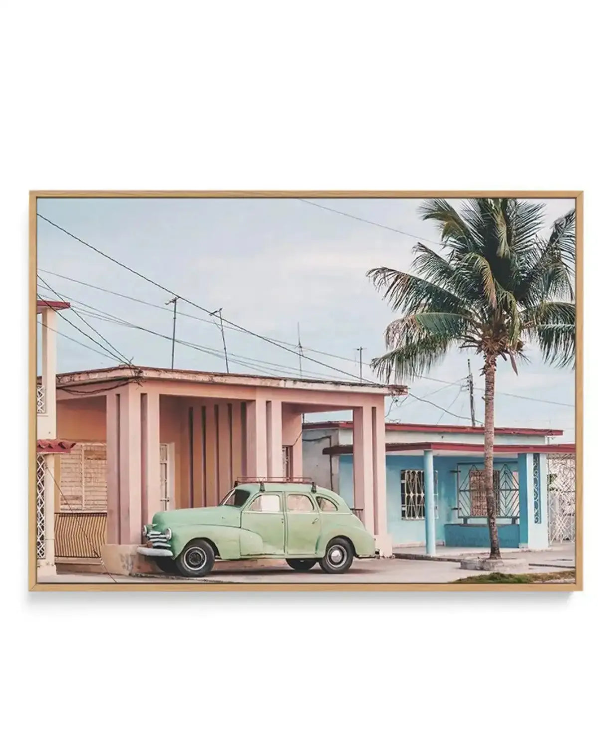 Framed canvas art print featuring a vintage-style photograph of a mint green 1950s automobile parked in front of pastel-colored buildings in warm dusty rose and turquoise blue tones. A tall palm tree stands beside the car on a street with power lines overhead and a clear sky. The canvas is mounted in a natural wood frame with a white border, creating a horizontal landscape composition that evokes a nostalgic Cuban street scene.